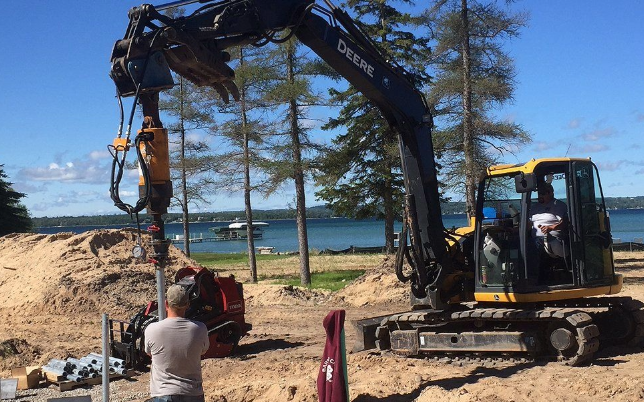 Helical pier installation in progress with an excavator and workers by the lakeside.